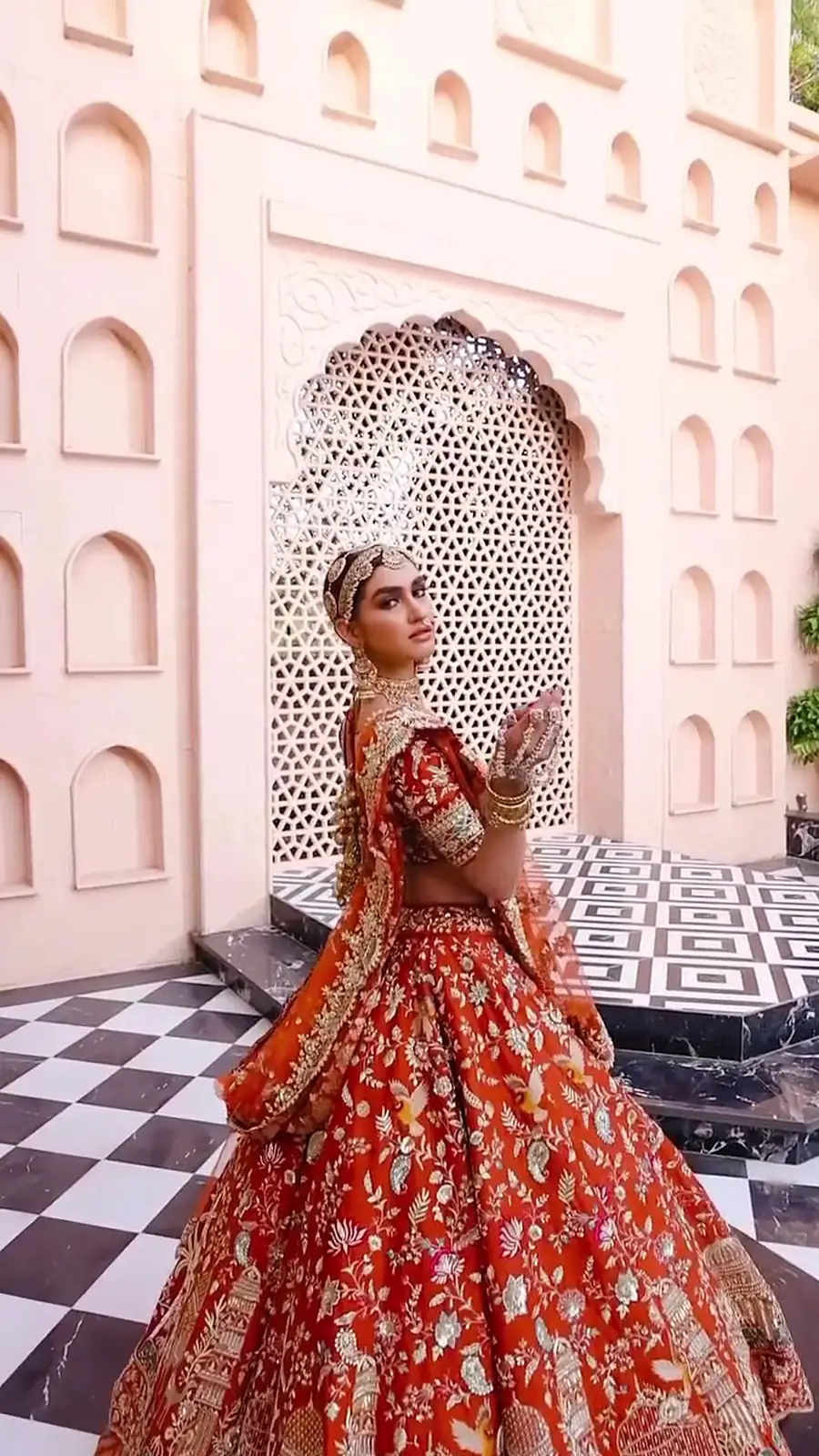 Bride in a coral bridal lehenga against a carved jaali archway